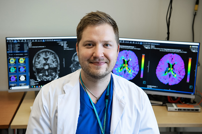 Valtteri Julkunen is sitting in front of a desk, there are several computer screens with brain imaging data behind him.