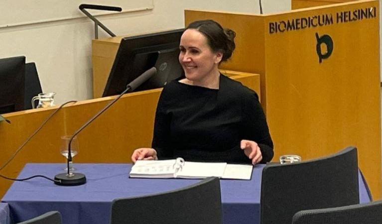 A woman in a black dress is seated at a desk in a lecture hall, smiling as she looks to her left.  A microphone, a folder and a glass of water are placed in front of her on the desk.