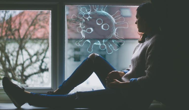 A woman is sitting on a windowsill, looking out of a window where you can see apartment buildings. A graphic of a large viral particle has been added to the image.