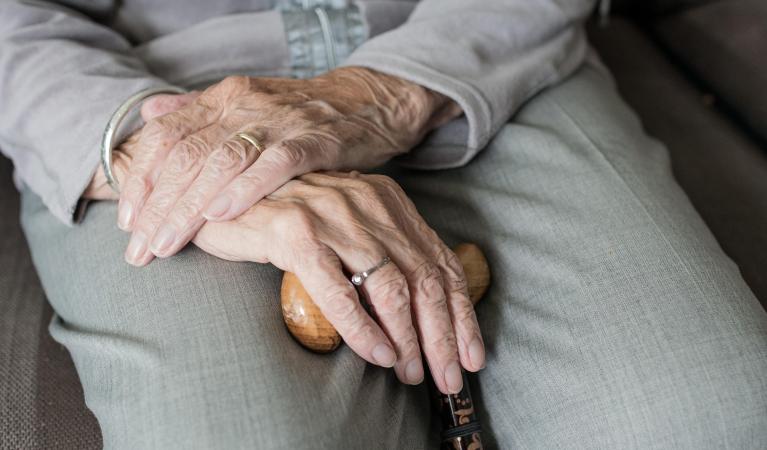 Close-up of an elderly person's hands resting on a wooden cane, with one hand gently placed over the other. 