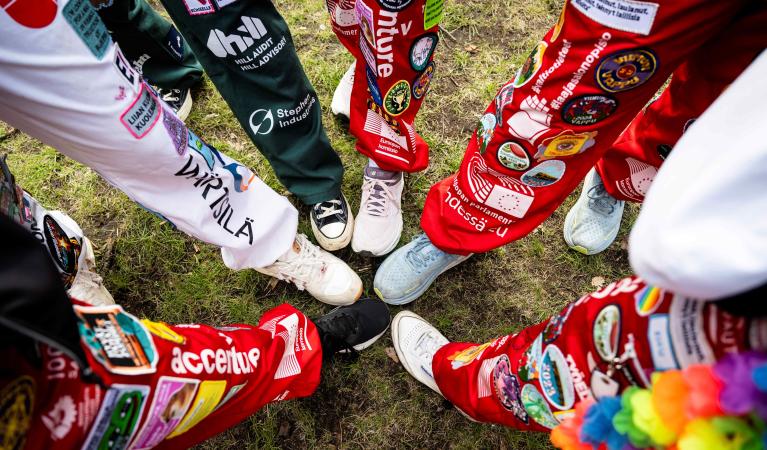 Eri värisiin opiskelijahaalareihin pukeutuneiden henkilöiden jalkoja ruohikolla.