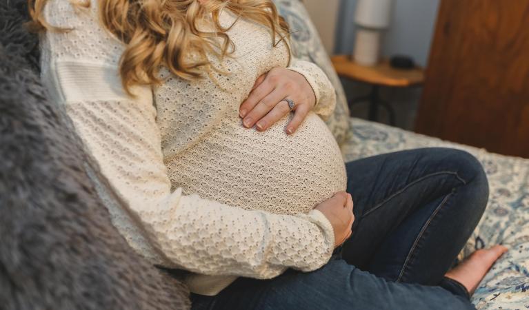 A woman is seated on a sofa wearing a light-colored knitted sweater and dark jeans, with both hands gently resting on a prominently rounded pregnant belly.