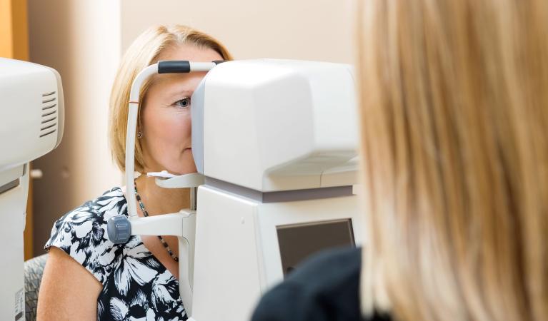 A woman with light hair looks into an eye examination device while a healthcare professional performs an eye test in a clinical setting.