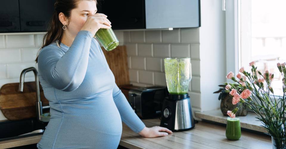A pregnant woman stands in the kitchen drinking a green smoothie.
