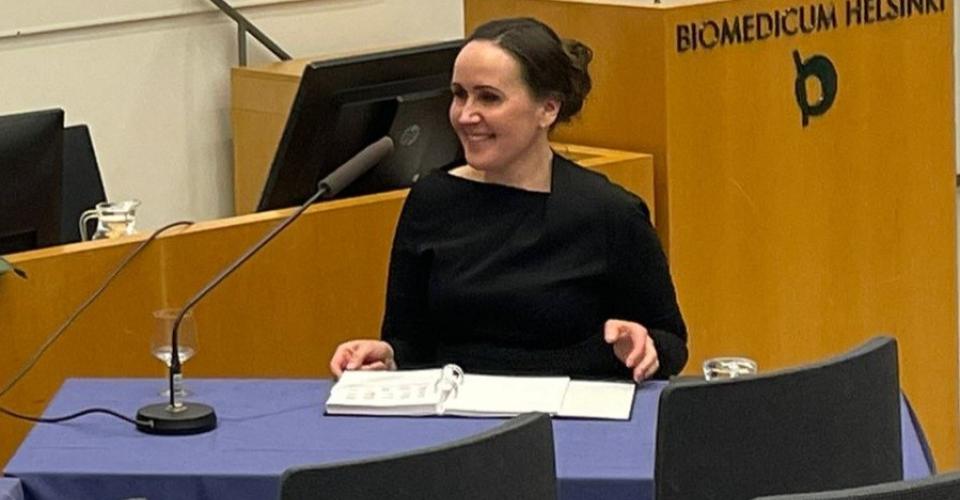 A woman in a black dress is seated at a desk in a lecture hall, smiling as she looks to her left.  A microphone, a folder and a glass of water are placed in front of her on the desk.