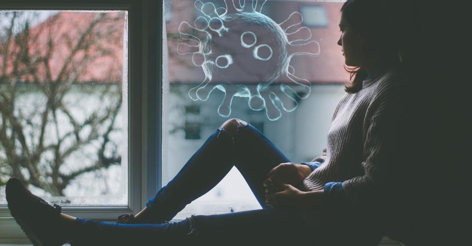 A woman is sitting on a windowsill, looking out of a window where you can see apartment buildings. A graphic of a large viral particle has been added to the image.