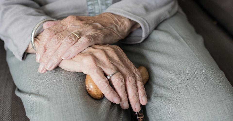 Close-up of an elderly person's hands resting on a wooden cane, with one hand gently placed over the other. 