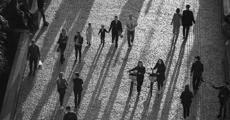 Black and white photo of people walking and cycling on a cobblestone street, casting long shadows in the late afternoon sunlight.