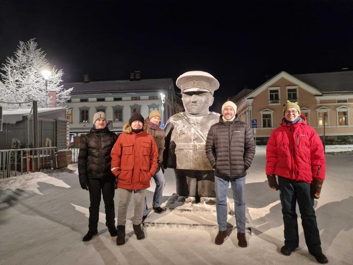 Viisi talvivaatteisiin pukeutunutta henkilöä seisoo lumessa yöllä Oulussa jykevän patsaan ympärillä. Patsas ja maa ovat kevyesti lumen peitossa, ja taustalla näkyy valaistuja rakennuksia.