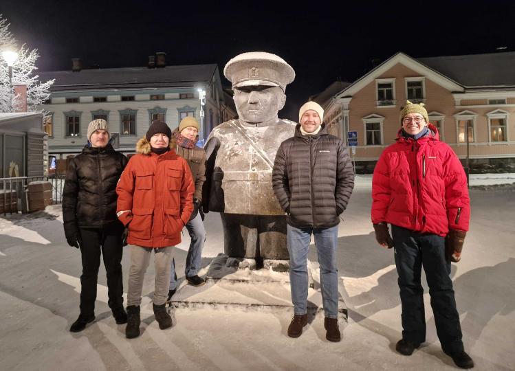 Five people in winter clothing stand in the snow at night in Oulu around a statue of a stout figure wearing a cap. The statue and ground are dusted with snow, and softly lit historic buildings line the background.
