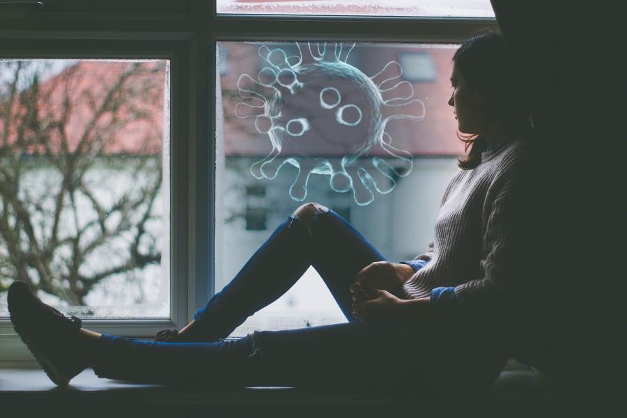 A woman is sitting on a windowsill, looking out of a window where you can see apartment buildings. A graphic of a large viral particle has been added to the image.
