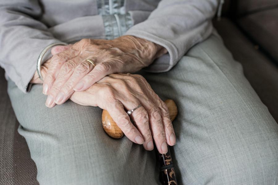 Close-up of an elderly person's hands resting on a wooden cane, with one hand gently placed over the other. 