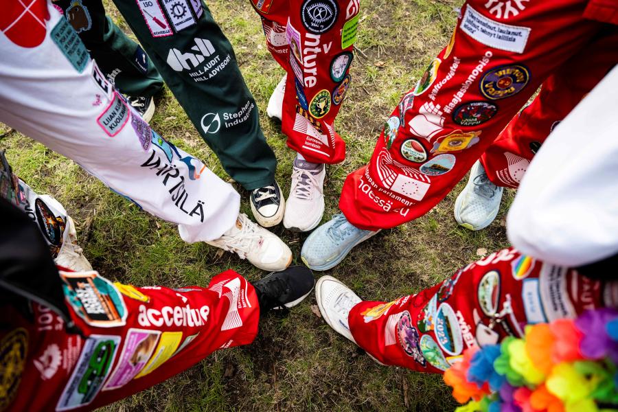 Eri värisiin opiskelijahaalareihin pukeutuneiden henkilöiden jalkoja ruohikolla.