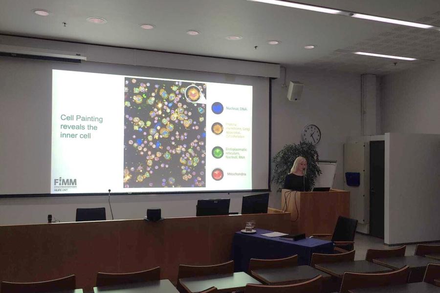 A doctoral researcher stands at a podium presenting her dissertation in a lecture hall, with a large screen displaying colorful cell painting images and labeled cellular components such as nucleus, mitochondria, and endoplasmic reticulum.