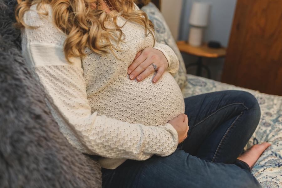 A woman is seated on a sofa wearing a light-colored knitted sweater and dark jeans, with both hands gently resting on a prominently rounded pregnant belly.
