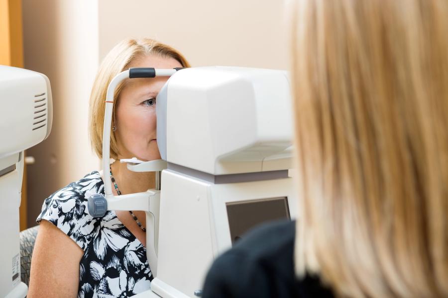A woman with light hair looks into an eye examination device while a healthcare professional performs an eye test in a clinical setting.